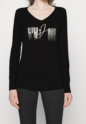 Frau trägt schwarzen Langarm-V-Neck-Oberteil mit silbernem LIU·JO-Logo aus kleinen Punkten und tropfenden Effekten auf der Vorderseite.
