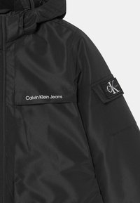 Černá vyp padded bunda s kapucí, s upevněním na zip, předními kapsami a logem Calvin Klein Jeans na hrudi a rukávu. Měkká textura.