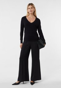 Schwarzer Strickpullover mit V-Ausschnitt, langen Ärmeln und strukturierten Bündchen, kombiniert mit weiten schwarzen Hosen und spitzen schwarzen High Heels.