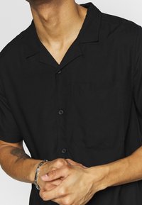 Lyhythihainen musta nappipaita, kevyt kangas, leirikaulus, yksittäinen rintatasku, pelkistetty malli, sileä pinta, näkyvät napit.