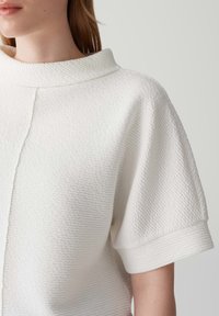 Top bianco testurizzato con colletto alto e ripiegato e maniche corte, caratterizzato da un sottile motivo a righe e cuciture pulite.