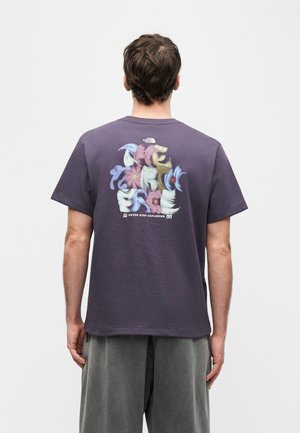 Moški, oblečen v temno vijolično majico s večbarvnimi abstraktnimi oblikami in napisom "Never Stop Exploring" na hrbtu, na belem ozadju.