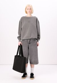 Felpa grigia con testo nero, pantaloni shorts di jeans grigi, borsa a tracolla nera e scarpe nere con suole beige. Il modello ha i capelli corti biondi.
