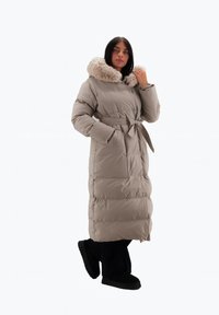 Abrigo largo acolchado beige con capucha forrada de piel, cintura ceñida y bolsillos laterales. Presenta textura acolchada y una silueta recta.