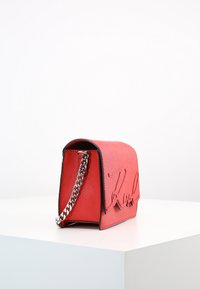 Borsa a spalla in pelle rossa con logo embosso, tracolla a catena argentata, forma rettangolare, texture liscia e rifiniture nere lungo i bordi.