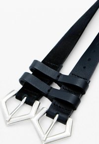 Cinturón de cuero negro con diseño de doble tira, hebilla triangular metálica y agujeros ajustables para variación de tamaño. Acabado de textura suave.