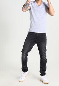 Světle šedé tričko s výstřihem do V, černé džíny s úzkým střihem a bílé tenisky s texturovaným povrchem a gumovou podrážkou. Viditelné tetování na paži.