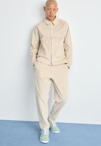 Beige, lätt bomullskappa med knäppning fram och sidofickor, tillsammans med matchande löst sittande byxor och gröna sneakers.