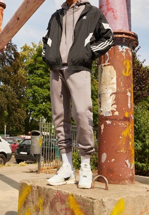 Personne portant une veste noire Adidas, un sweat à capuche gris et un pantalon de jogging, des chaussettes blanches et des baskets, debout sur un bloc de béton près d'un poteau rouillé en extérieur.