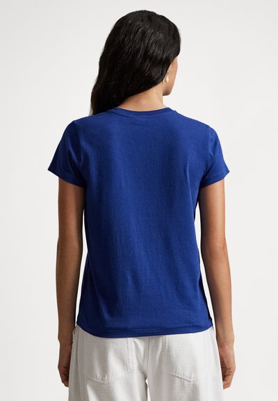 Kvinne med langt svart hår iført en ensfarget blå kortermet t-skjorte og hvite bukser, stående med ryggen til mot en ensfarget bakgrunn.