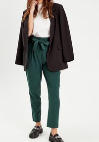 Femme portant un pantalon taille haute en papier vert, une blouse blanche, un blazer noir drapé sur les épaules, et des mocassins noirs avec des détails en chaîne argentée.