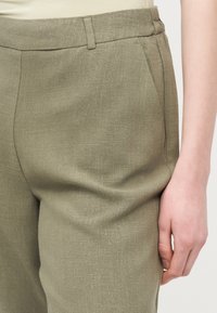 Κοντινό πλάνο ενός ατόμου που φοράει ελιά-πράσινο παντελόνι με υφή και πλαϊνές τσέπες, εμφανίζοντας μέρος του χεριού που ακουμπάει δίπλα στο μηρό.