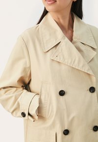 Beige dobbeltradet frakke med store sorte knapper, bred fornærmelseskrave og sidelommer. Glat stof med diskret glans.