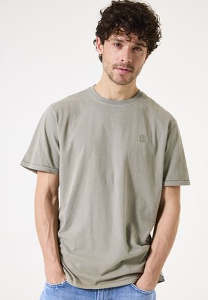 Camiseta de algodón gris con mangas cortas, cuello redondo y un pequeño logo en el pecho. La tela parece suave y tiene un corte relajado.