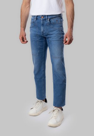 Uomo che indossa jeans blu medio a gamba dritta con grandi tasche frontali, sneakers bianche e t-shirt bianca, in piedi su uno sfondo grigio.