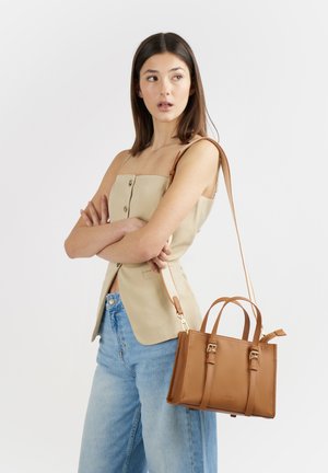 Donna con top beige senza maniche e jeans blu che porta una borsa di pelle marrone chiaro con tracolla, guardando di lato su sfondo bianco.