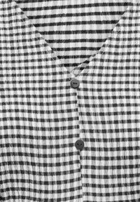 Melnbalta raibs faktūrēts audums ar divām redzamām melnām pogām un V veida izgriezuma detaļu.