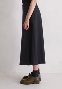 Jupe noire, longueur genou avec une texture lisse et une taille élastique, associée à des chaussures marron à lacets et des chaussettes sombres. Vue de profil.