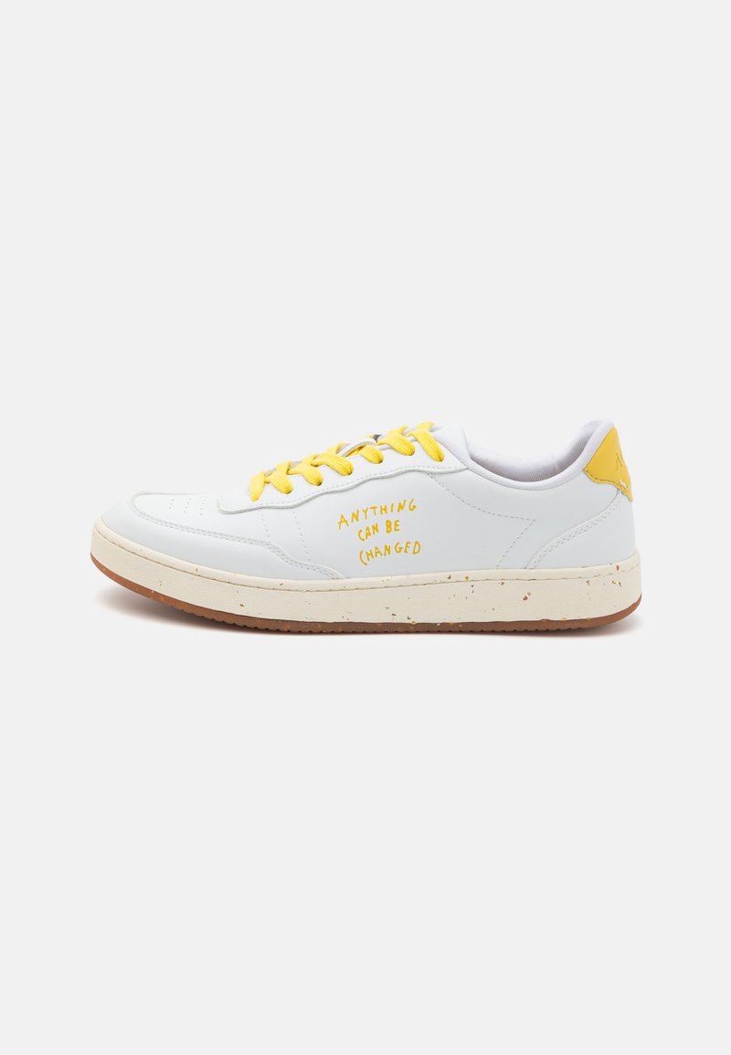 White sneaker with yellow laces and accent, featuring a textured midsole with speckled details. Text "ANYTHING CAN BE (changed)" on the side.