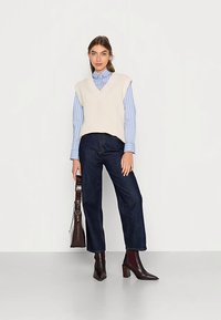 Gilet lavorato a maglia color crema con scollo a V, abbinato a una camicia con bottoni a strisce azzurre e jeans a gamba larga di colore blu scuro. Stivaletti marroni alla caviglia e borsa a tracolla.