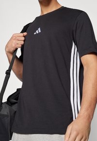 Svart bomull t-shirt med rund hals, korta ärmar och tre vita vertikala ränder på varje sida. Inkluderar en liten logotyp på bröstet.