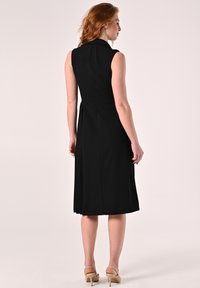 Zwarte mouwloze jurk met kraag, voorzien van een aansluitend lijfje en een uitlopende rok. Soepele textuur, tot op de knie, met een rits aan de achterkant.