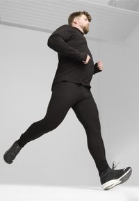 Schwarzes Langarmoberteil kombiniert mit schwarzen engen Leggings, beide aus dehnbarem Material. Das Modell trägt schwarze Sportschuhe mit weißen Akzenten.