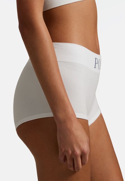 Kobieta w białej, bezszwowej bieliźnie z wysokim stanem, z napisem "POLO" na gumce, pokazana z profilu na jednolitym tle.