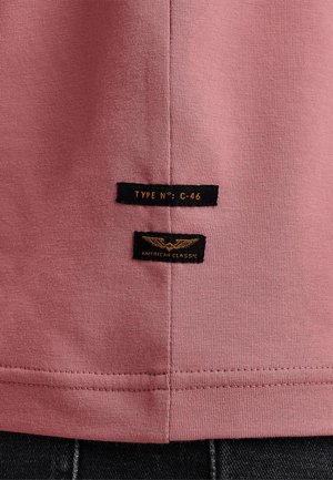 Rosa Baumwoll-T-Shirt mit glatter Textur, ausgestattet mit zwei schwarzen Etiketten: "TYPE No: C-46" und "AMERICAN CLASSIC" mit goldenen Akzenten.