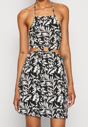 Robe dos nu noire et blanche avec motif de feuilles et découpe frontale en forme d'anneau, portée par une personne tatouée les bras le long du corps.
