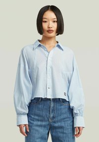 Camicetta azzurra chiara accorciata con bottoni frontali, maniche lunghe a sbuffo e una texture sottile; abbinata a jeans di denim.