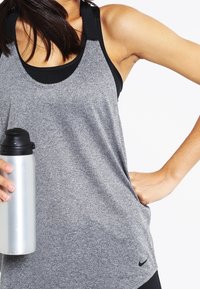 Grå träningslinne med svarta accenter, gjort av texturerat tyg. En silverfärgad vattenflaska hålls i ena handen. Nike-logotyp synlig.