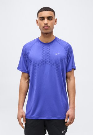 Ung mand stående iført en lilla Nike sportsskjorte med korte ærmer og sorte shorts mod en ensfarvet lys baggrund.