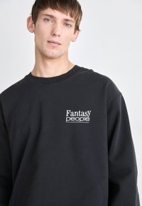 Schwarzer Baumwollpullover mit Rundhalsausschnitt und langen Ärmeln. Auf der linken Brust befindet sich der weiße Schriftzug "Fantasy people". Sanfte Textur, lässige Passform.