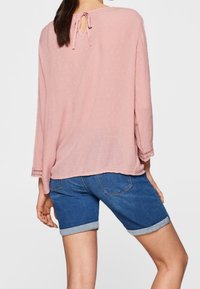 Hellrosa Bluse mit dreiviertel Ärmeln, strukturiertem Stoff, Rückverschluss mit Schleife, kombiniert mit Jeans-Shorts mit umgeschlagenen Säumen.