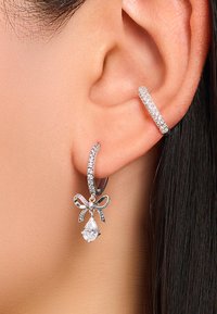 Silberne Ohrringe mit einem Ringdesign, versehen mit glitzernden Steinen und einer dekorativen Schleife. Ein tropfenförmiger Kristall hängt von der Schleife.