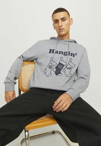 Sivi dukserica s natpisom "Hangin'" i tri dekorativne čarape. Opusti fit, elastične manžetne i prednja džep u obliku kengura, uparen s crnim hlačama.