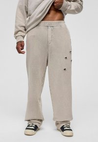Beige sweatpants met een relaxte pasvorm, met zichtbare stiksels en drie beschadigde details op het rechterbeen. Draagt het met zwarte sneakers.
