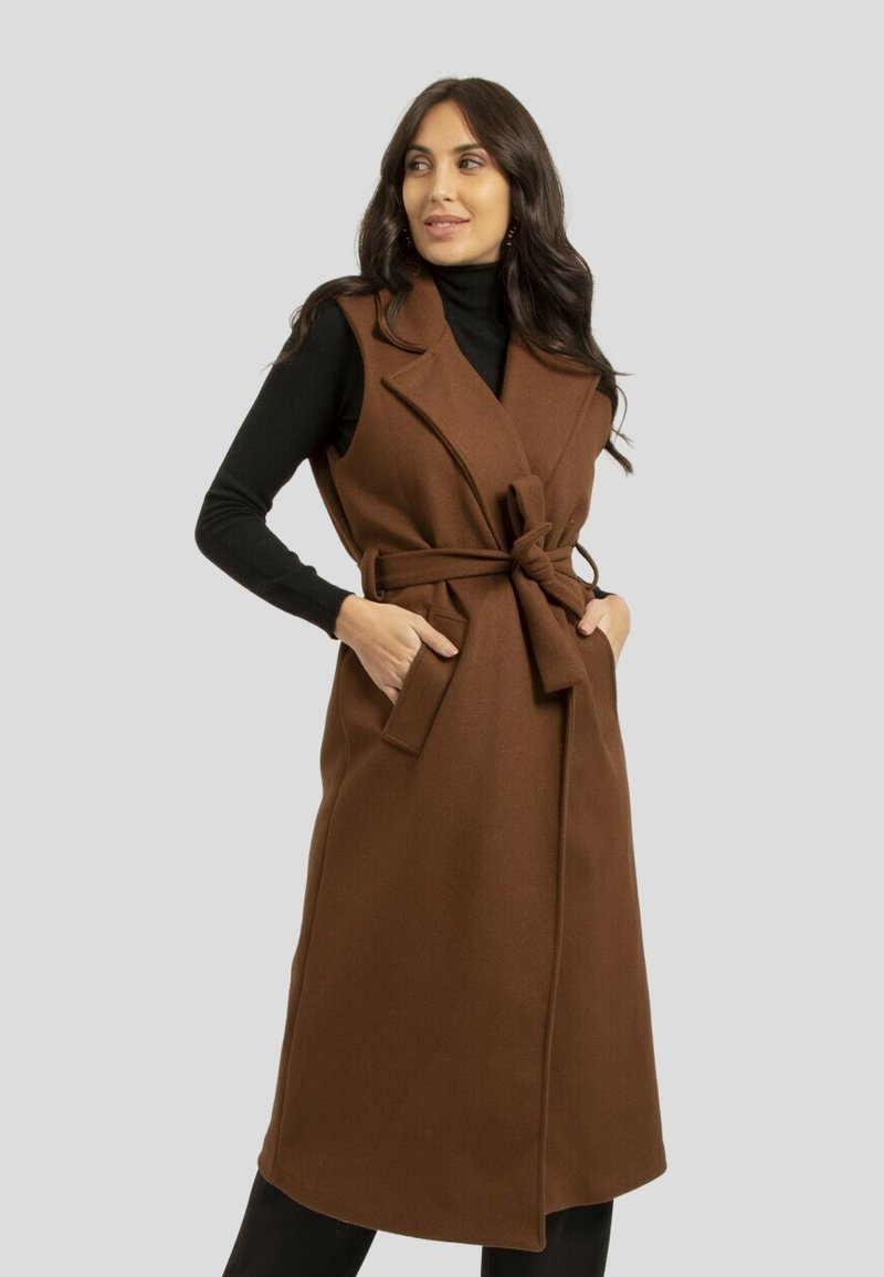 Manteau sans manches marron avec une taille ceinturée, des poches latérales et un col en v, fabriqué à partir d'un tissu texturé, superposé sur un col roulé noir.