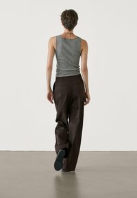 Harmaa ribbattu tanktoppi yhdistettynä löysiin ruskeisiin housuihin, joissa on sivutaskut, nähtynä takaa vaalealla lattialla yksinkertaisessa taustassa.