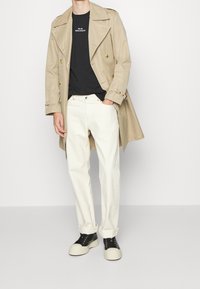 Beżowy płaszcz trench na czarnym t-shircie, kremowych dżinsach i czarno-białych sneakersach. Wyróżniające szczegóły to podwójne guziki i podwinięte mankiety.