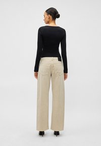 Γυναίκα με κότσο, φορούσε μαύρο τοπ με μακριά μανίκια, μπεζ παντελόνι φαρδύ στα μπατζάκια και μαύρα ψηλοτάκουνα παπούτσια, στέκεται με την πλάτη προς το μέρος μας.