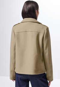 Beige villasekoitteinen takki, jossa on rakennettu kaulus, olkaepauletit ja siisti takapintakuvio. Esitetty tummien farkkujen kanssa, sileä tekstuuri.