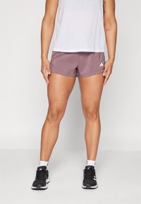 Žena stojící v mauve atletických šortkách, bílém bezrukávovém topu, bílých ponožkách a černých teniskách adidas na jednobarevném pozadí.