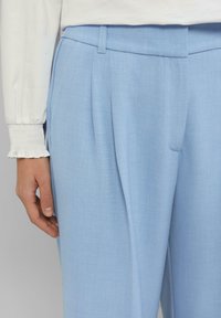 Lichtblauwe op maat gemaakte broeken met een gladde textuur, voorzien van plooien en een plat tailleband, weergegeven naast een witte longsleeve top.