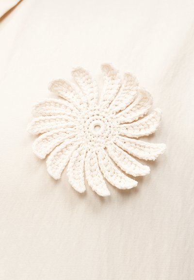 Πλεκτή λευκή ανθική εφαρμογή με στρωμένα πέταλα σε μπεζ υφασμάτινο φόντο, αναδεικνύοντας υφασμένο νήμα και περίπλοκο σχέδιο.