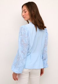 Light blue blouse featuring solid fabric at the back and sheer lace embroidered sleeves with floral patterns. Structured, flared sleeves.