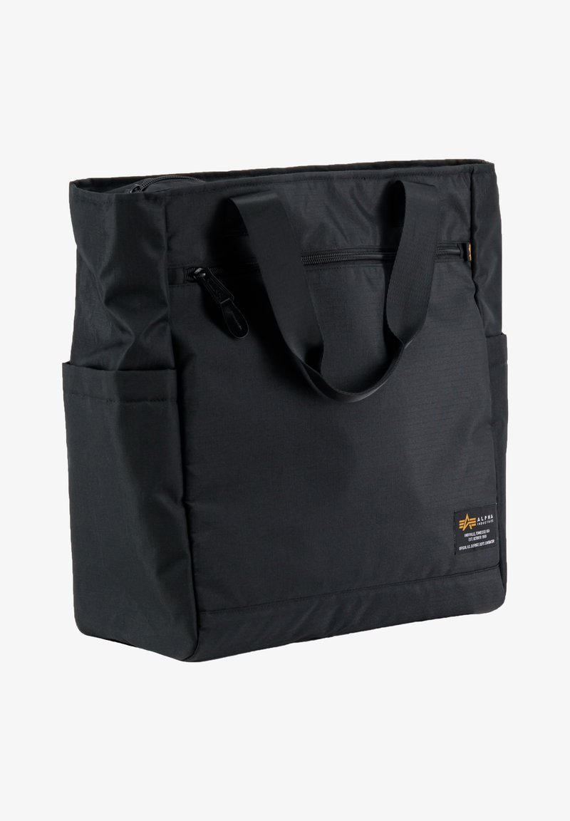 Sac fourre-tout en tissu noir avec un haut zippé, deux poignées et des poches latérales. Présente un petit logo et une surface texturée.