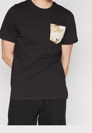 Homme portant un t-shirt noir avec une poche camouflage marron clair et beige étiquetée "Tommy Jeans" sur la poitrine gauche.