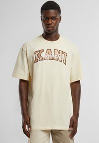 Cream cotton t-shirt with short sleeves featuring a large brown graphic text "KANI" and "Originato" in white cursive across the chest.